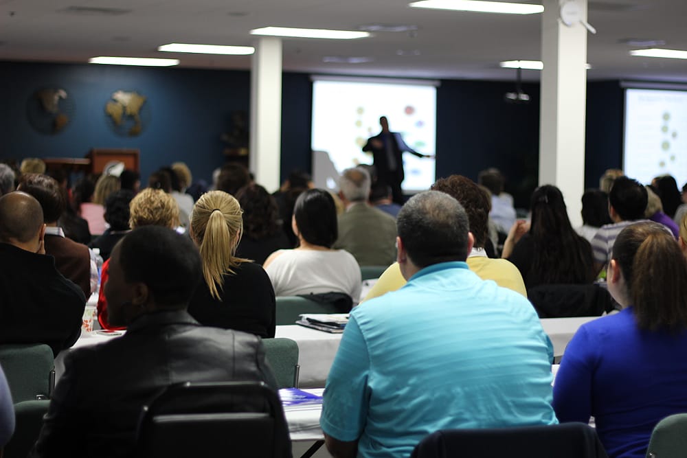 A group of professionals seated in a conference room, attentively watching a speaker at a podium during an MGE seminar.