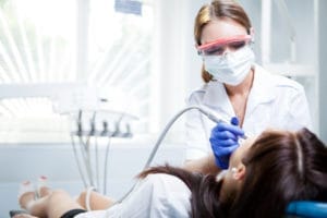 Dental hygienist in protective gear cleaning a patient's teeth with dental tools.