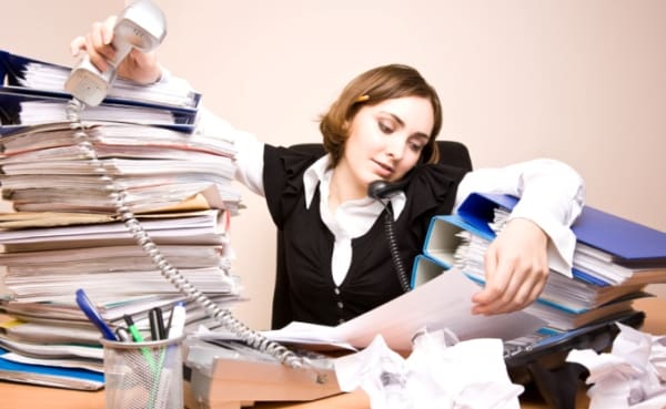 A woman appearing overwhelmed while working at a cluttered desk with phone and papers.