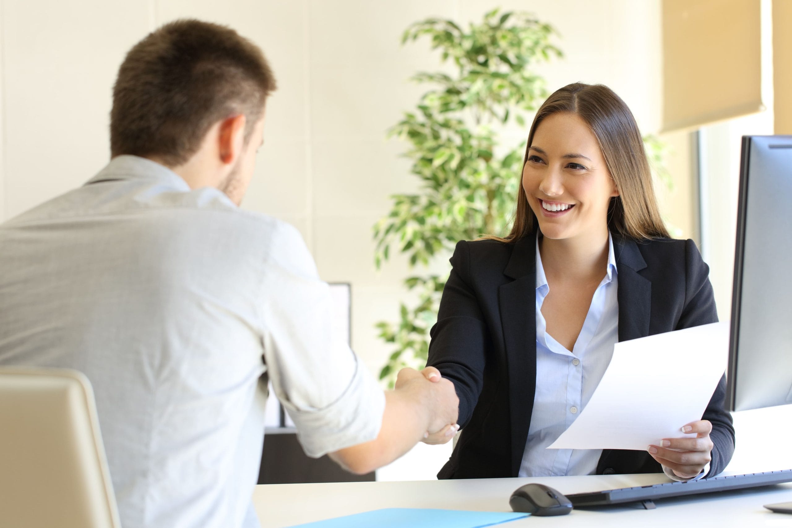 A female holding a paper across the table shaking hands with a male in a job interview.