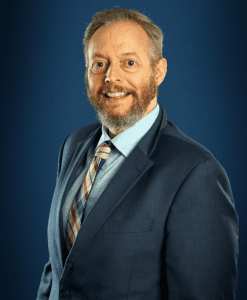 A head and shoulders portrait of a middle-aged man with short dark hair, wearing a dark suit and tie, smiling directly at the camera.