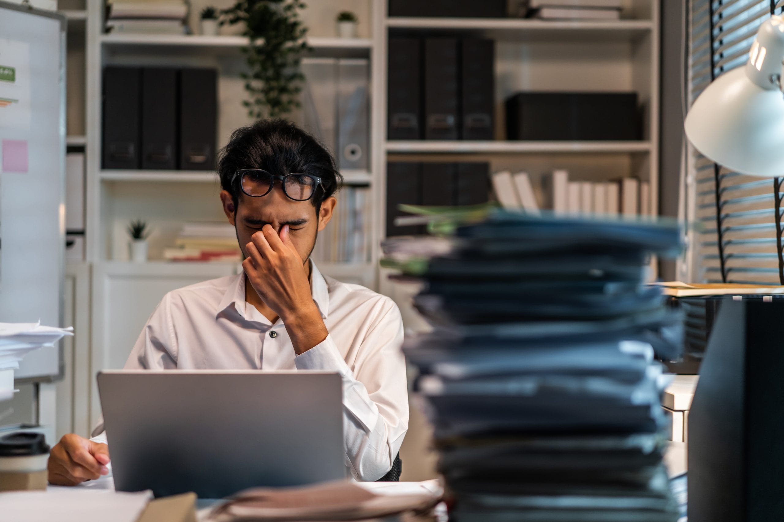 A man behind a desk with piles of paperwork, looking overwhelmed.