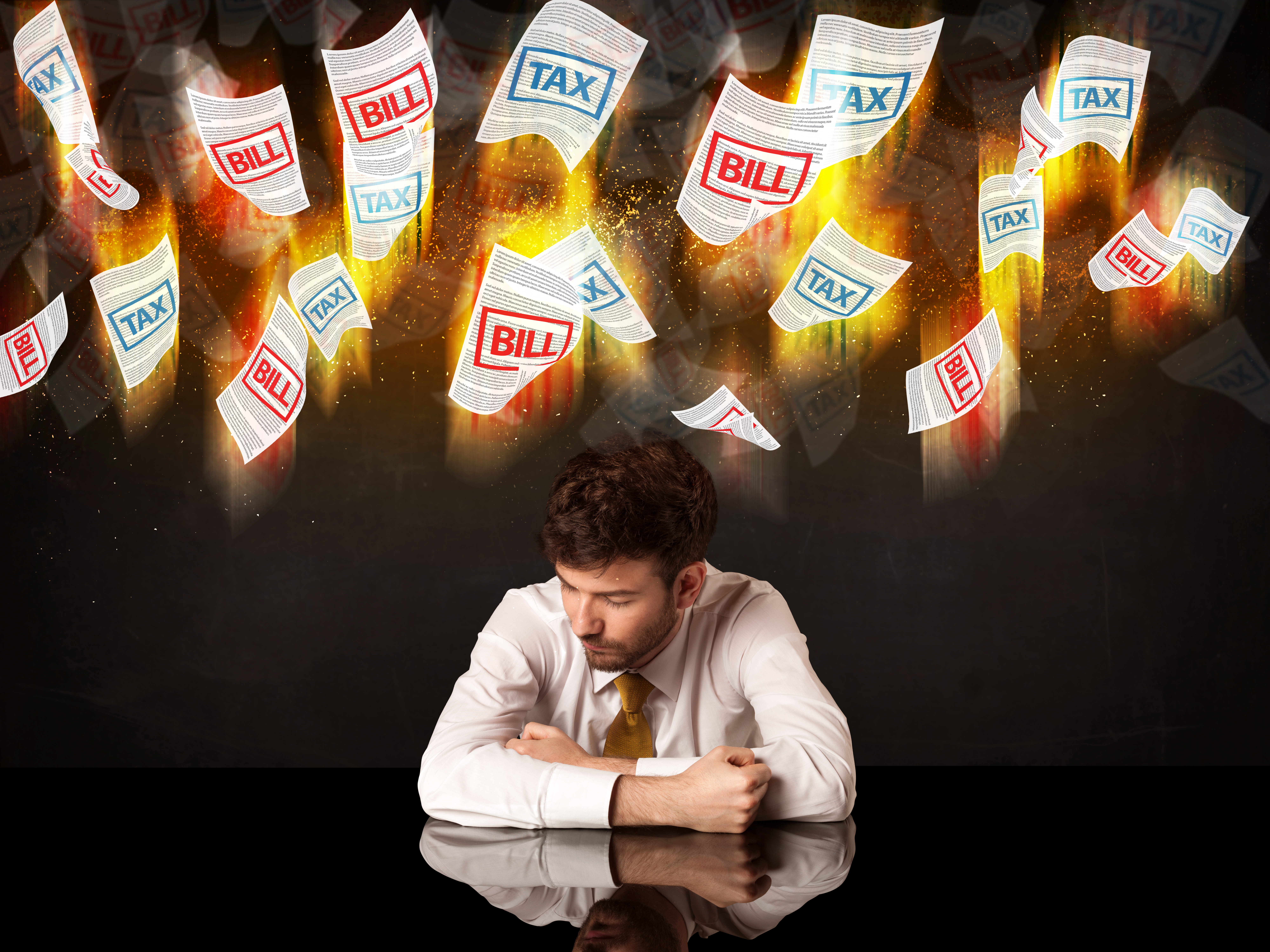 A male sitting with his head close to the table and pieces of flaming paper with the words bills and tax flying above his head.